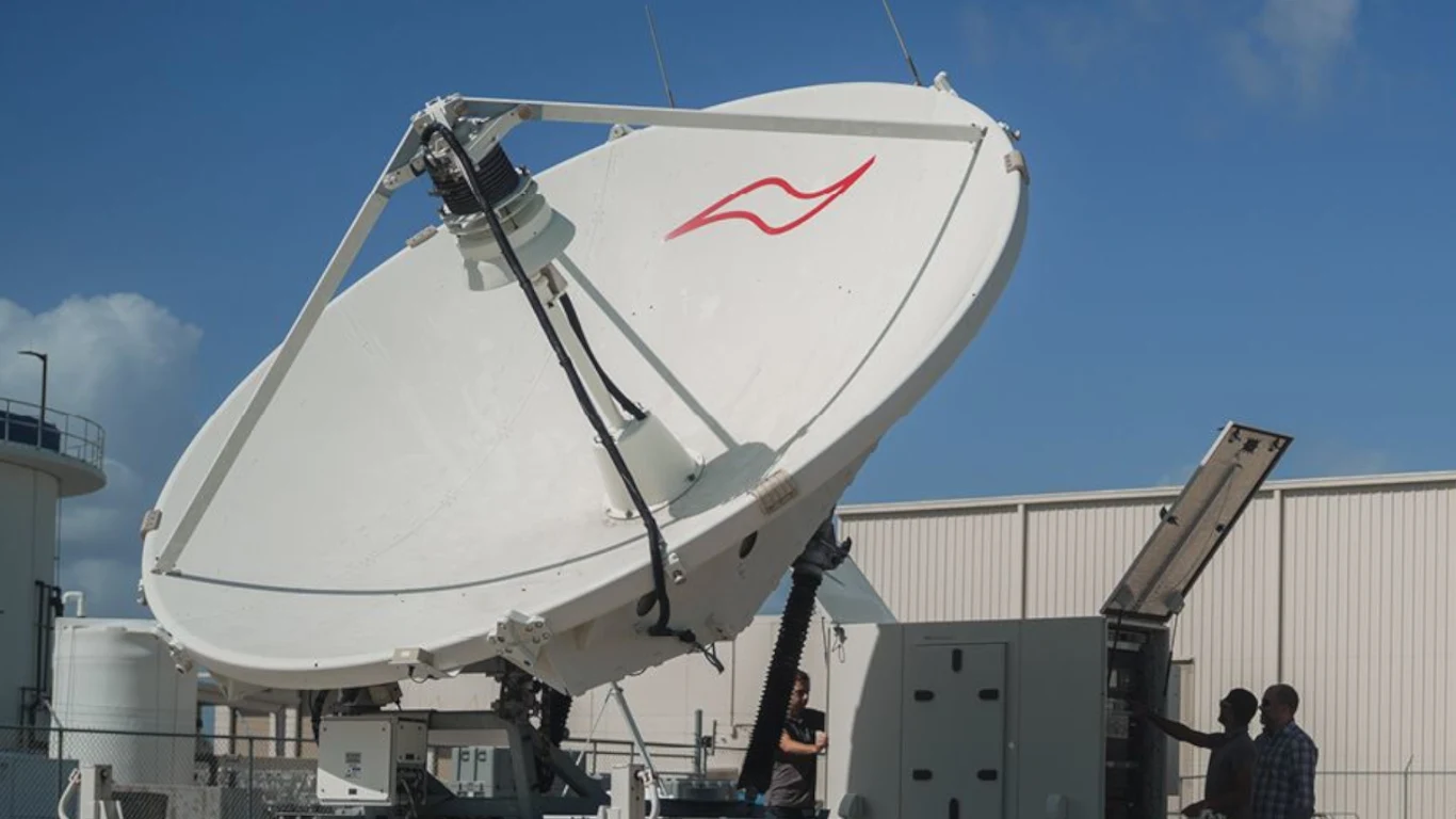 Duża antena satelitarna skierowana w stronę nieba, z boku znajduje się obudowa techniczna obsługiwana przez dwie osoby. W tle widać budynki przemysłowe, a niebo jest bezchmurne.