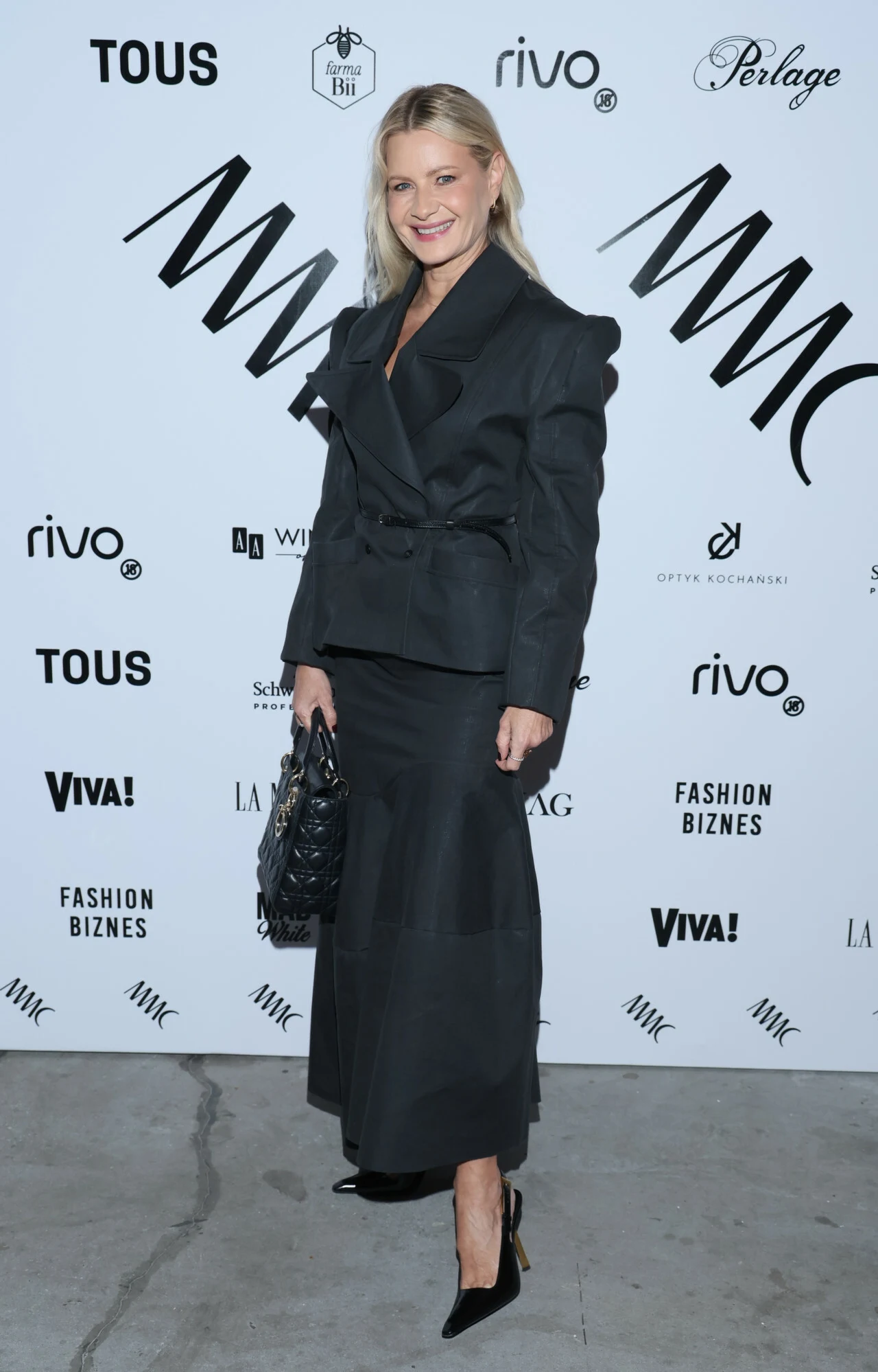 Kobieta w eleganckim, ciemnym garniturze z długą spódnicą i marynarką stoi na tle ścianki z logotypami marek, trzymając torebkę i uśmiechając się do aparatu.