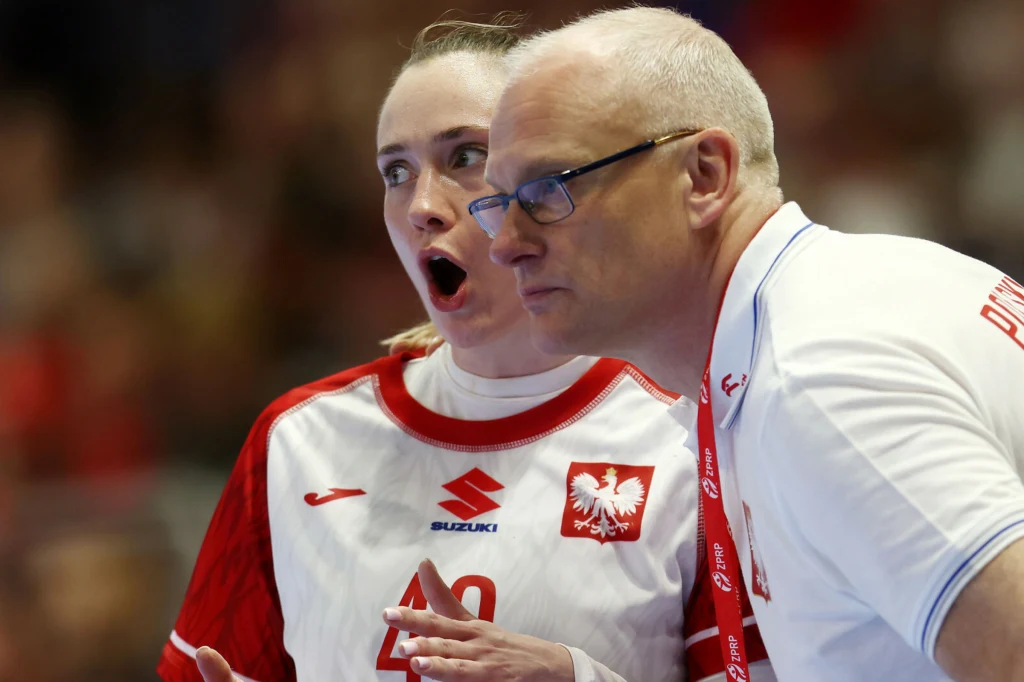 Zawodniczka polskiej reprezentacji piłki ręcznej w trakcie rozmowy z trenerem, oboje w sportowych strojach z polskim godłem, na tle rozmytego tłumu.