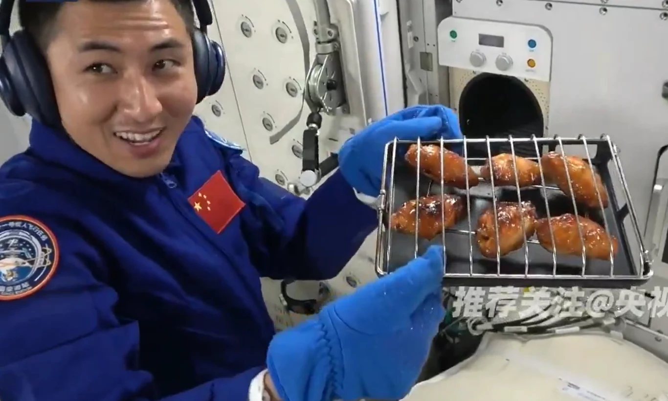 Kosmiczne skrzydełka wyglądają apetycznie. Uśmiechy chińskich astronautów mówią same za siebie Astronauta w niebieskim skafandrze trzyma tacę z pieczonymi skrzydełkami kurczaka w wnętrzu statku kosmicznego. Na rękach ma niebieskie rękawice ochronne, a w tle widoczne są elementy wyposażenia technicznego.