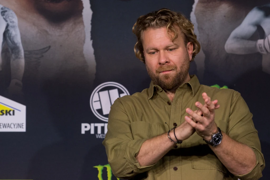 Mężczyzna z brodą i falującymi, blond włosami stoi w jasnej koszuli na tle plakatów reklamowych i logo. Składa dłonie jakby klaskał lub pocierał dłonie, ze spokojnym wyrazem twarzy. W tle widoczny fragment rysunku lub zdjęcia przedstawiającego postaci ...