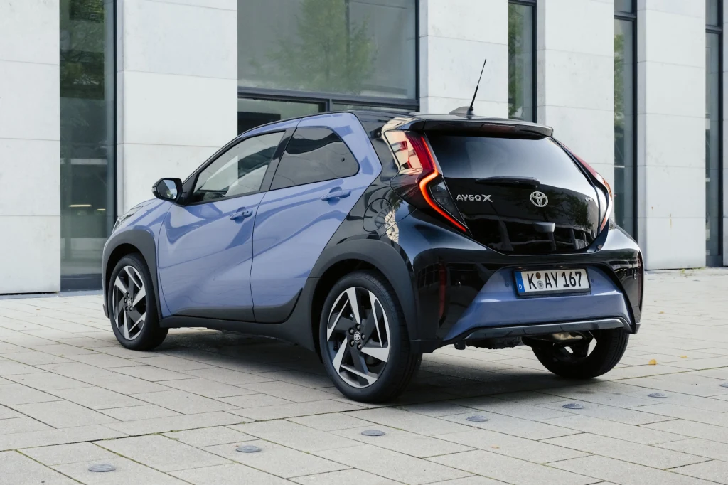 Toyota Aygo X z tyłu praktycznie się nie zmieniła Nowoczesny samochód osobowy Toyota Aygo X w kolorze niebiesko-czarnym, zaparkowany na tle szklanego budynku z jasną elewacją. Wyrazista stylistyka tylnych świateł LED i dynamiczne linie nadwozia podkreślają kompaktową sylwetkę auta.