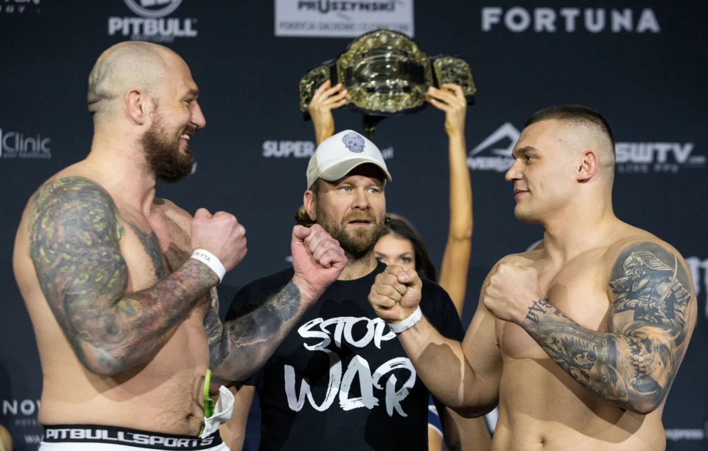 Dwóch umięśnionych, wytatuowanych mężczyzn stoi naprzeciwko siebie w pozycji do walki, pomiędzy nimi znajduje się mężczyzna w czapce z daszkiem ubrany w koszulkę z napisem 'Stop War', w tle widoczny jest pas mistrzowski trzymany przez kobietę oraz rekl...