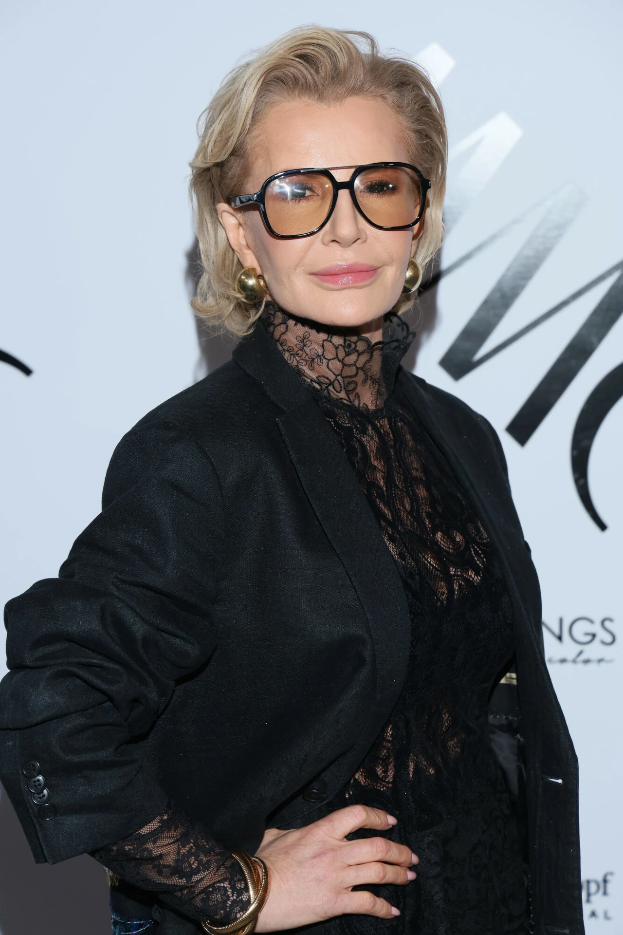 Joanna Racewicz w modnej stylizacji Kobieta w okularach o jasnych włosach ubrana w elegancki czarny koronkowy strój i czarną marynarkę, pozuje na tle z białym logo i napisem. Jej wyraz twarzy jest poważny, a postawa pewna siebie.