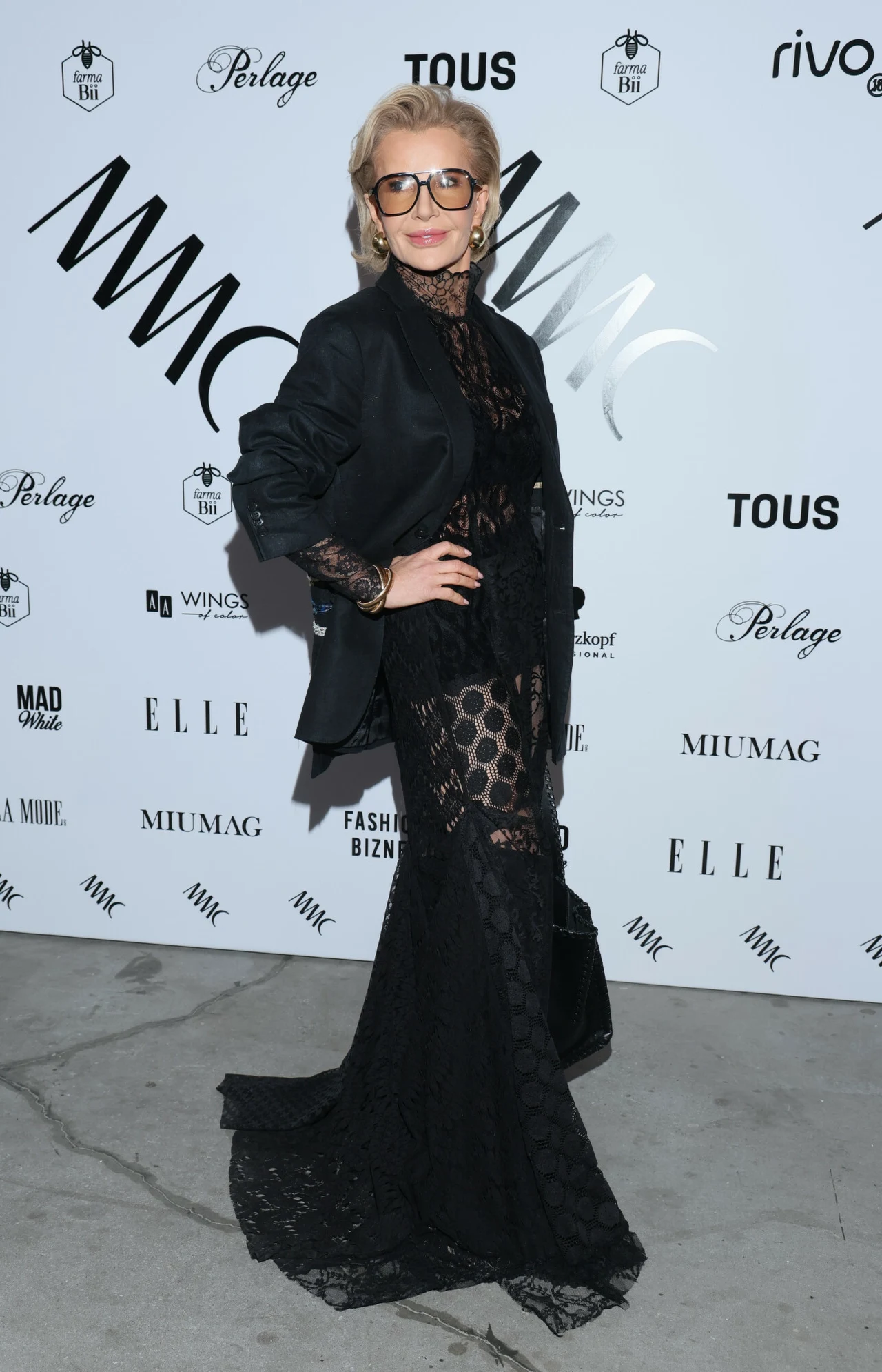 Joanna Racewicz lubi bawić się modą Elegancka kobieta w długiej, czarnej, koronkowej sukni i marynarce, pozująca na tle ściany z logotypami sponsorów, ubrana w okulary i trzymająca czarną torebkę.
