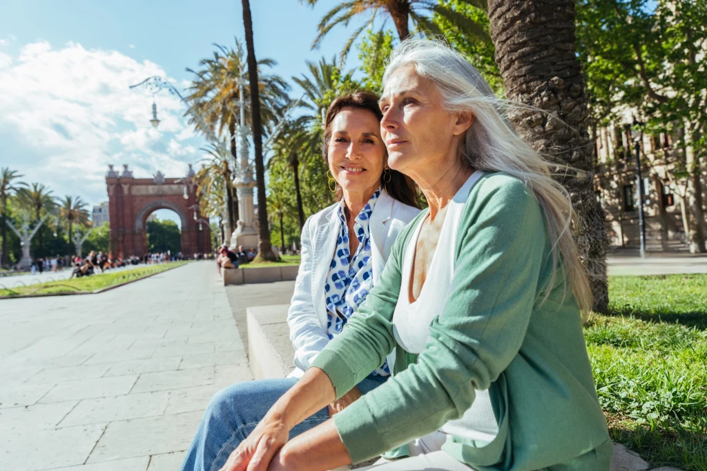 Dwie starsze kobiety siedzące na schodach w słoneczny dzień, otoczone palmami i zielenią, w tle widoczny jest łuk triumfalny i miejskie alejki.