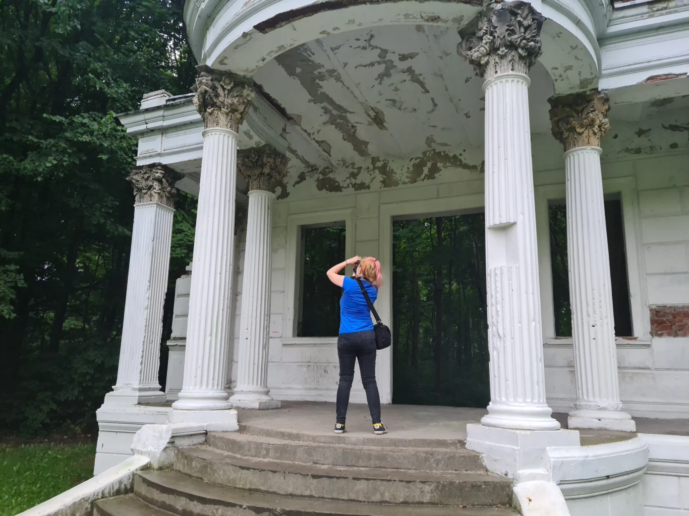 Woj. opolskie jest pełne atrakcji - także tych mniej oczywistych. Kobieta w niebieskiej koszulce fotografuje zaniedbaną kolumnadę z białymi, uszkodzonymi kolumnami, na tle zielonych drzew. Architektura budynku wykazuje widoczne ślady zniszczenia i łuszczącej się farby.