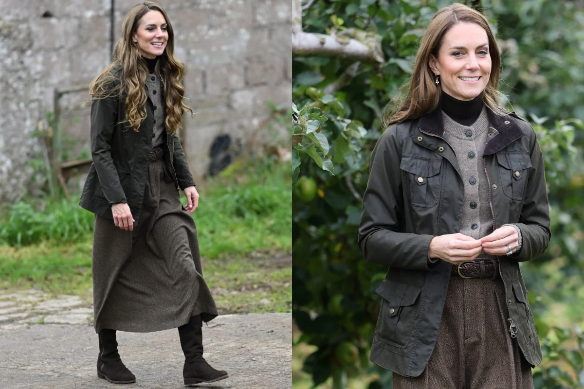 Ulubiona kurtka na jesień księżnej Kate Kobieta o długich, falowanych włosach ubrana w brązową sukienkę, granatową kurtkę oraz ciemne buty, stoi na tle zieleni i starego, kamiennego muru. Uśmiechnięta i zrelaksowana postawa, naturalne otoczenie.