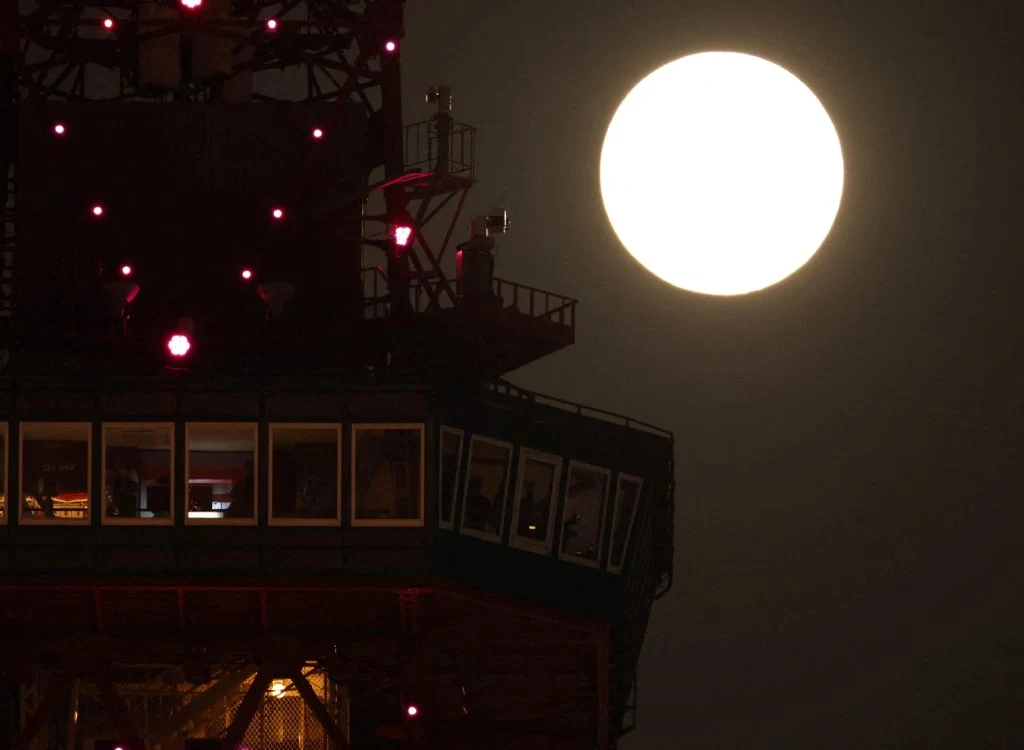 Wieża z oświetleniem nocnym, na tle pełnej, jasno świecącej tarczy księżyca.