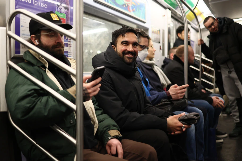Mamdani w nowojorskim metrze Grupa mężczyzn siedzi na ławce w wagonie metra, niektórzy korzystają z telefonów, jeden z nich uśmiecha się szeroko. W tle widać innych pasażerów oraz charakterystyczne wnętrze metra.