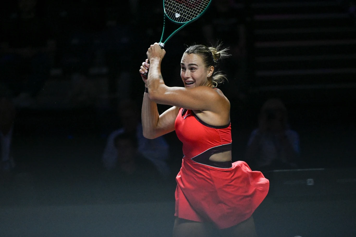 Aryna Sabalenka podczas WTA Finals 2025 Tenisistka w czerwonym stroju wykonuje zamach rakietą tenisową w trakcie meczu, skupiona na akcji, na tle ciemnego, rozmytego stadionu.