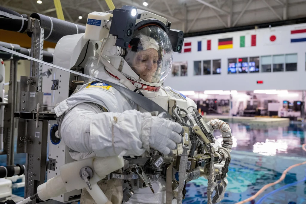 Astronauta w pełnym skafandrze kosmicznym wykonuje czynności techniczne przy specjalistycznym sprzęcie w dużym basenie treningowym, w tle widoczne są flagi międzynarodowe i sprzęt laboratorium.