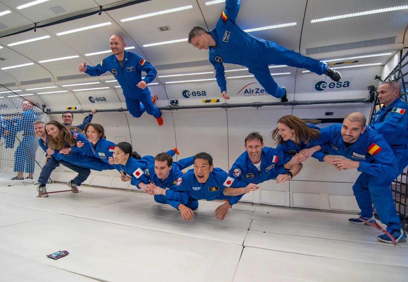 Grupa osób ubranych w niebieskie kombinezony z naszywkami krajowymi i logo ESA unosi się w stanie nieważkości w specjalnie przystosowanym wnętrzu samolotu, jedna osoba trzyma się siatki, uczestnicy wyglądają na podekscytowanych i skupionych.