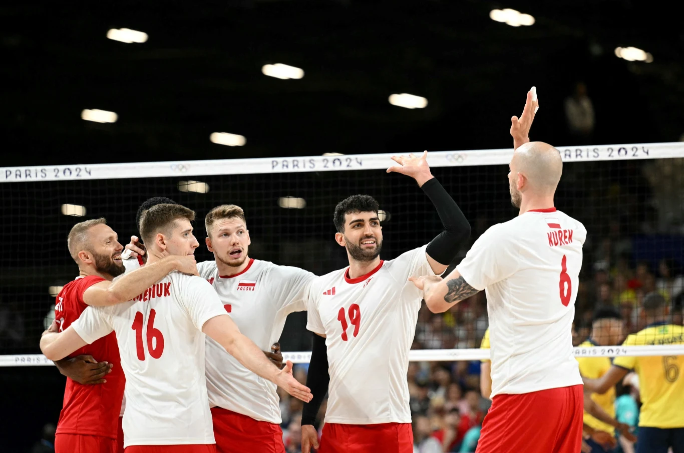 Grupa siatkarzy w białych koszulkach z numerami świętuje zdobycie punktu na boisku podczas meczu, w tle widoczni kibice oraz siatka z napisem Paris 2024.