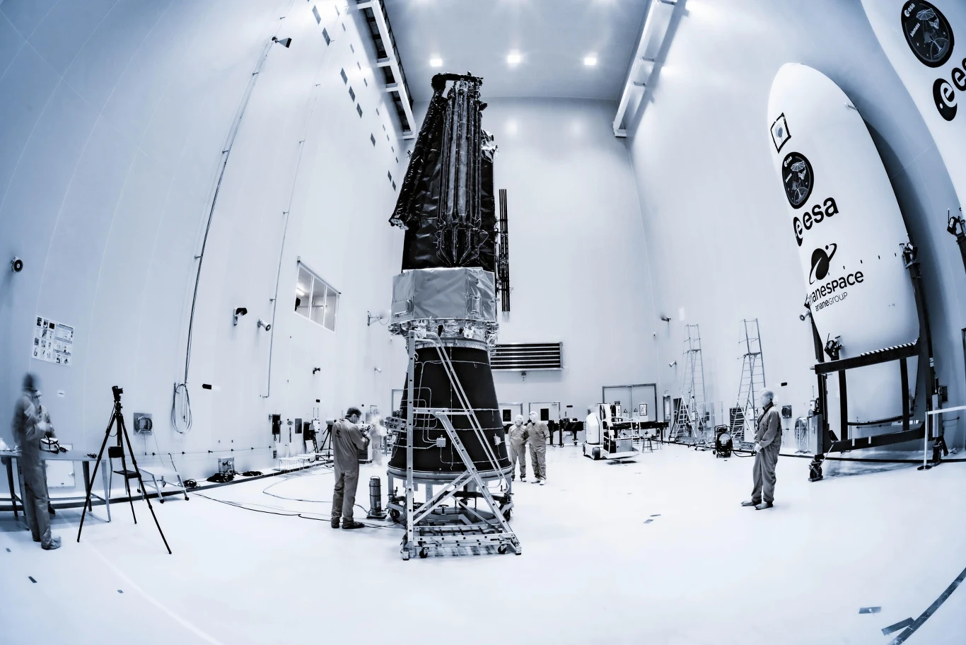 Grupa pracowników technicznych pracuje przy dużej konstrukcji statku kosmicznego w jasnej, sterylnej hali montażowej, obok elementów oznaczonych logotypami ESA i Arianespace.