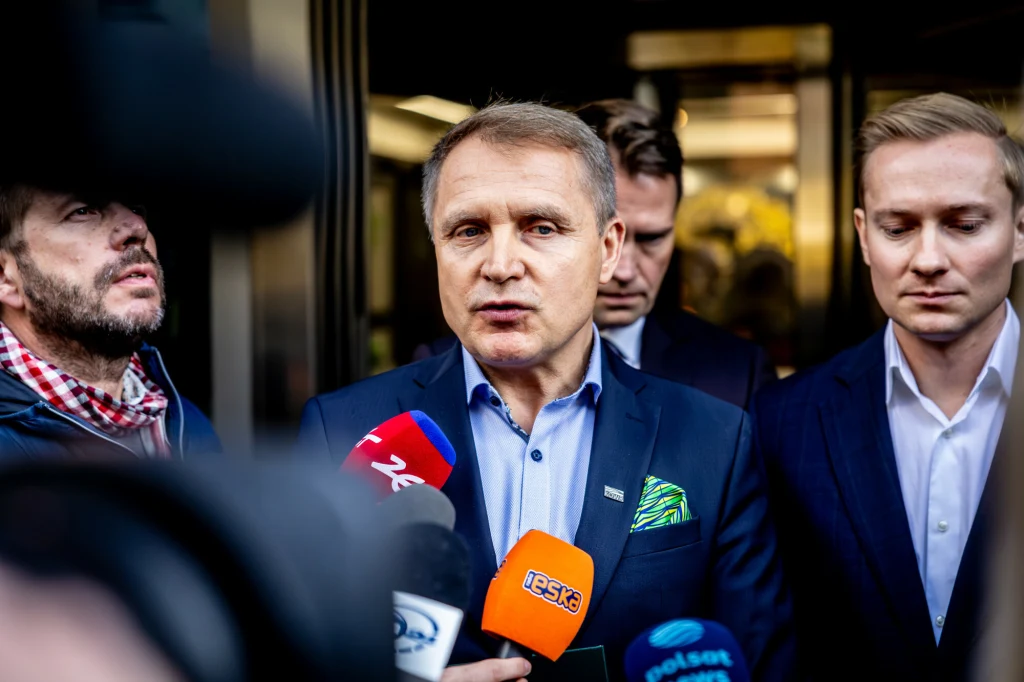 Mężczyzna w garniturze udziela wywiadu mediom, otoczony przez innych mężczyzn i mikrofony kilku stacji telewizyjnych, w tle niewyraźne wnętrze budynku.