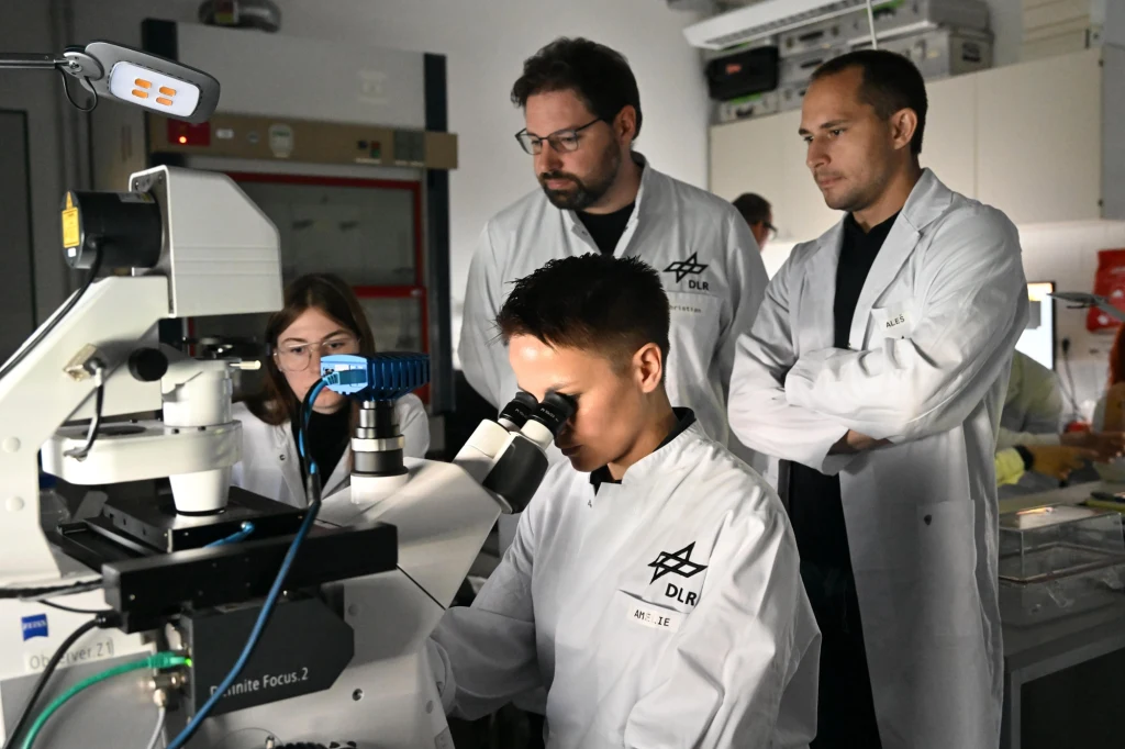 Grupa naukowców w białych fartuchach laboratoryjnych obserwująca młodego chłopca patrzącego przez mikroskop w nowoczesnym laboratorium, w tle specjalistyczny sprzęt oraz elementy wyposażenia pracowni badawczej.