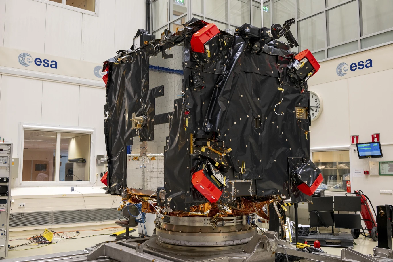 Wnętrze nowoczesnego laboratorium ESA z dużym satelitą lub sondą kosmiczną umieszczoną na specjalnym stanowisku testowym, urządzenie otoczone licznymi elementami technicznymi, światłem oraz narzędziami serwisowymi.