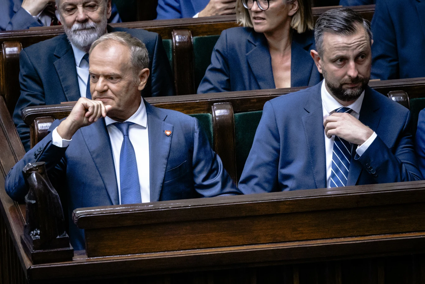 Dwóch mężczyzn w garniturach siedzi obok siebie na ławkach sejmowych, w tle inni uczestnicy posiedzenia, wszyscy w formalnych strojach, atmosfera poważna, oficjalna.