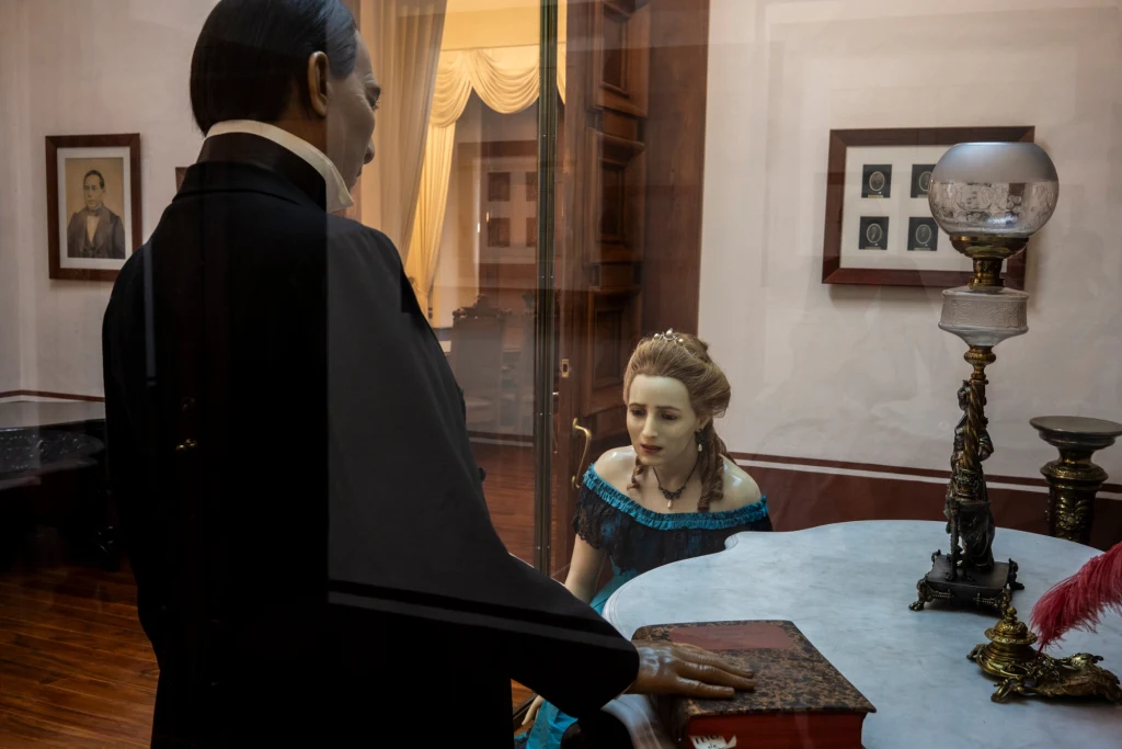 Woskowe figury historycznych postaci w eleganckim gabinecie, mężczyzna pochyla się nad siedzącą kobietą przy stole, wokół klasyczne meble, lampa naftowa i oprawione zdjęcia na ścianach.