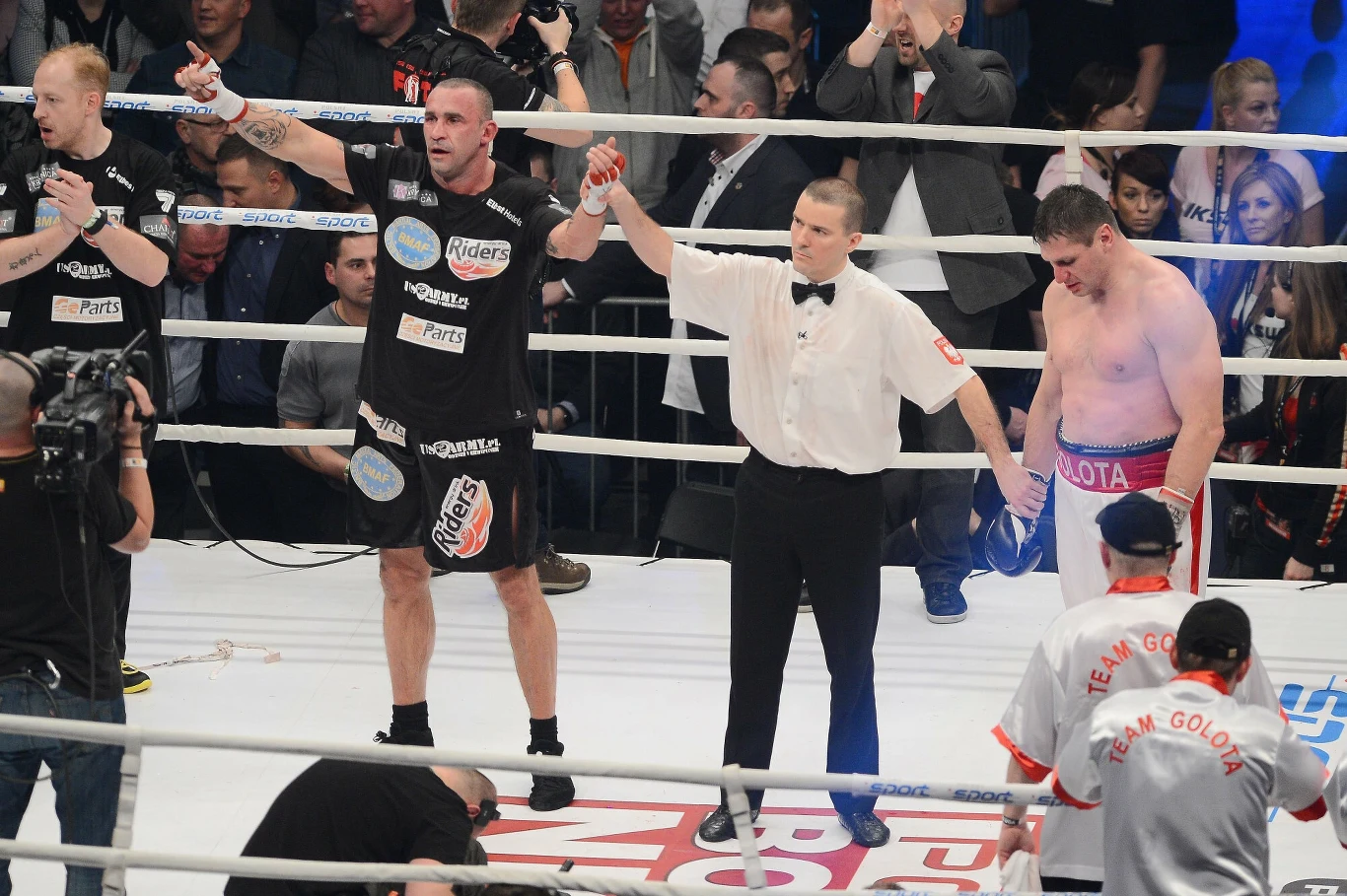 Zwycięzca walki bokserskiej z uniesioną ręką stoi obok sędziego, który także trzyma rękę przegranego zawodnika, podczas gdy wokół w ringu znajdują się trenerzy i obsługa. Publiczność obserwuje wydarzenie.