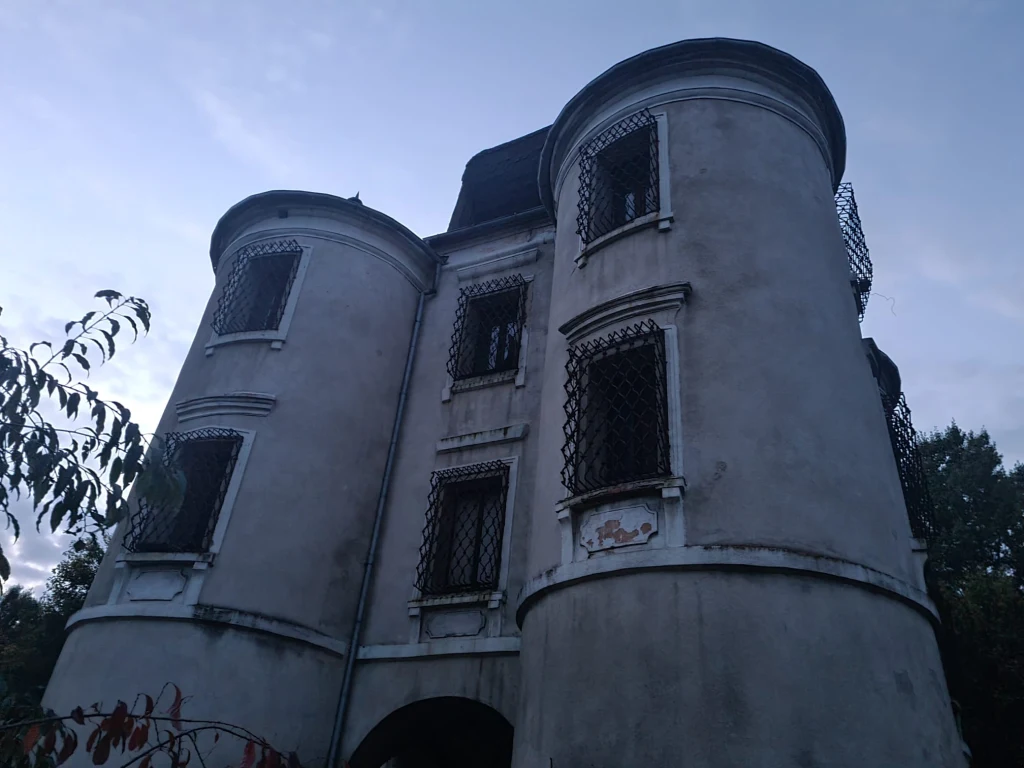 Belweder stanowił pawilon ogrodowy, a w wieżyczkach znajdowały się pomieszczenia dla gości. Stara, opuszczona rezydencja z oknami zabezpieczonymi kratami, o dwóch okrągłych wieżach, fasada budynku miejscami zniszczona, wokół drzewa i gęsta roślinność, niebo zachmurzone.