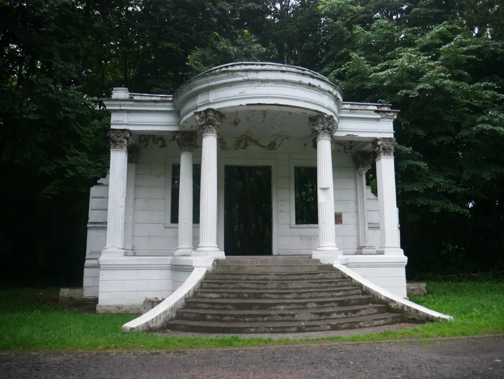 Zwiedzając Polskę, często kierujemy się tylko do popularnych miejsc. Warto to zmienić i lepiej poznać historię! Biała rotunda z czterema kolumnami i schodami prowadzącymi do wejścia, nieco zniszczona, otoczona gęstymi zielonymi drzewami.