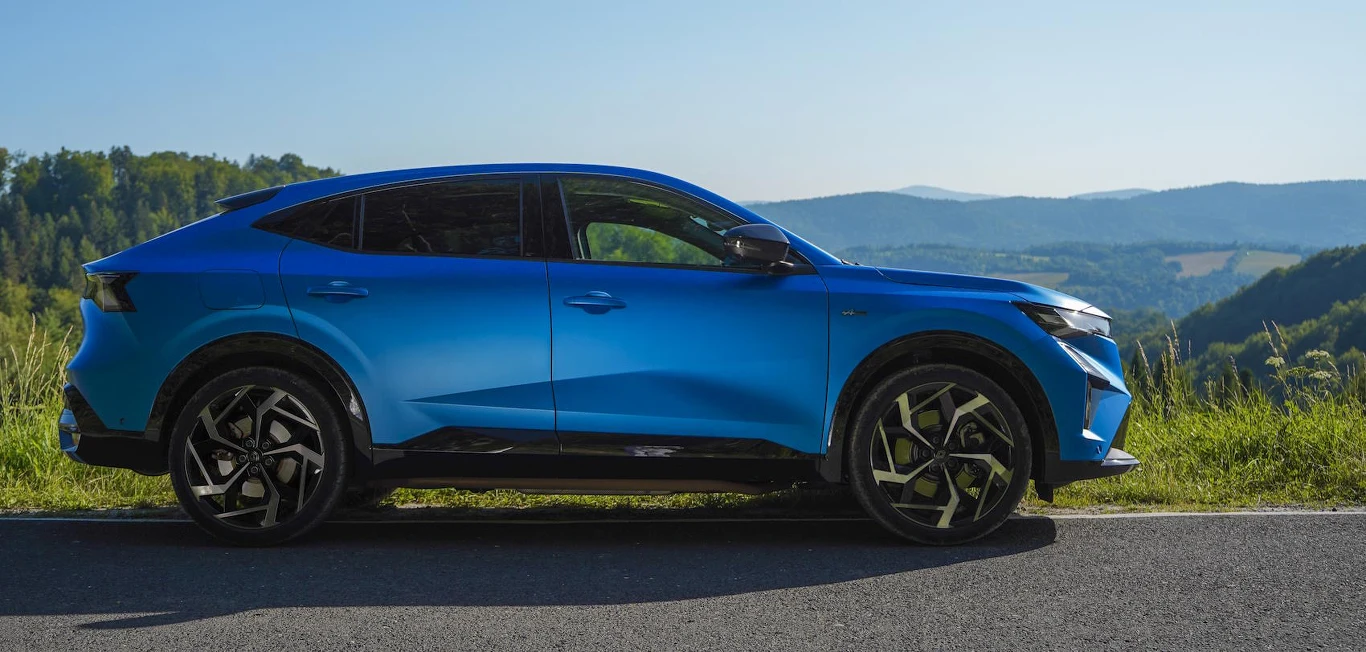 Rafale to pełnoprawny SUV segmentu C Nowoczesny niebieski SUV zaparkowany przy drodze, w tle rozciągają się zielone wzgórza i góry oraz czyste niebo.