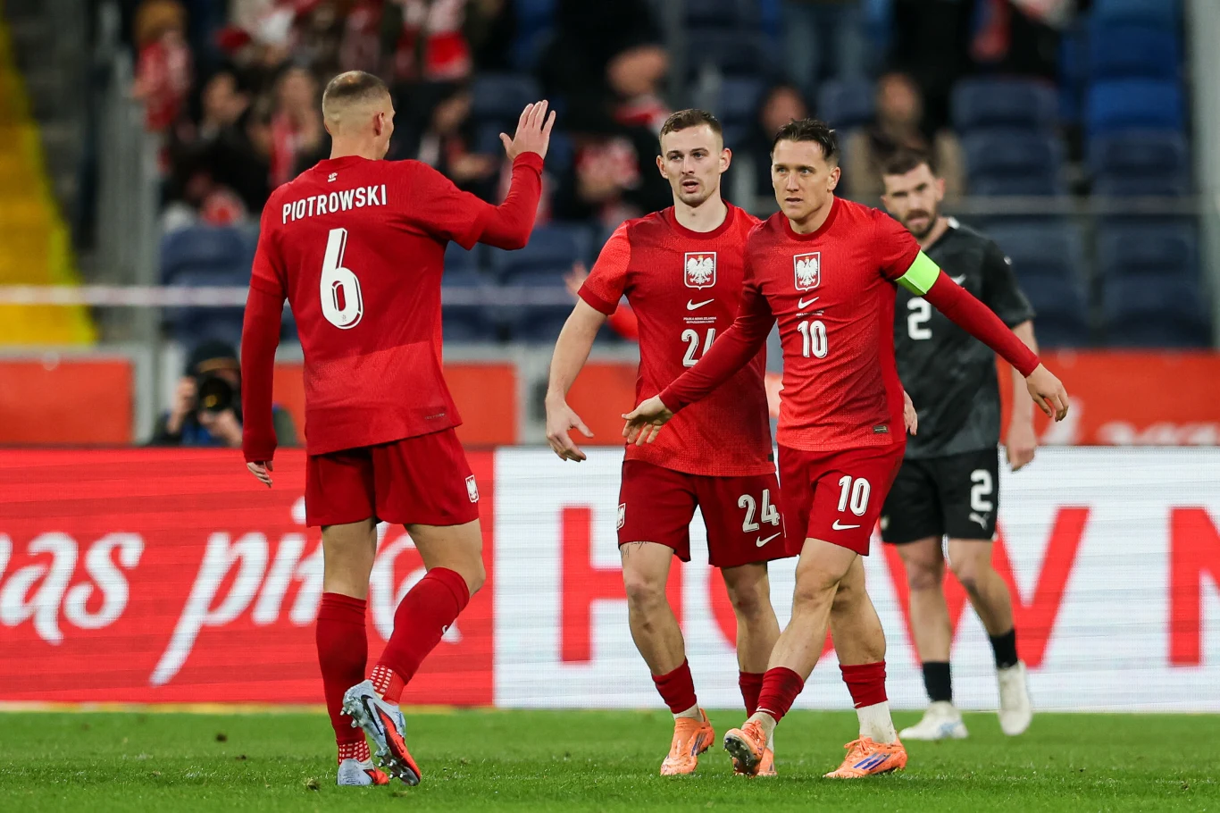 Zawodnicy reprezentacji Polski w piłce nożnej świętują akcję podczas meczu, ubrani w czerwone stroje z widocznymi numerami i nazwiskami, na tle stadionu oraz kibiców.