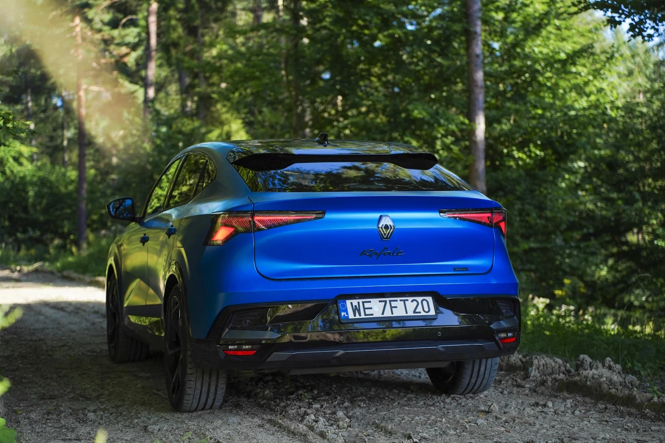 Auto zaskakuje pojemnym bagażnikiem – wystarczy, żeby spakować czteroosobową rodzinę na wakacyjny wyjazd Nowoczesny, niebieski samochód Renault Austral o sportowej sylwetce stoi na leśnej drodze otoczonej zielonymi drzewami. Z tyłu widoczna jest charakterystyczna linia świateł LED oraz polska rejestracja.