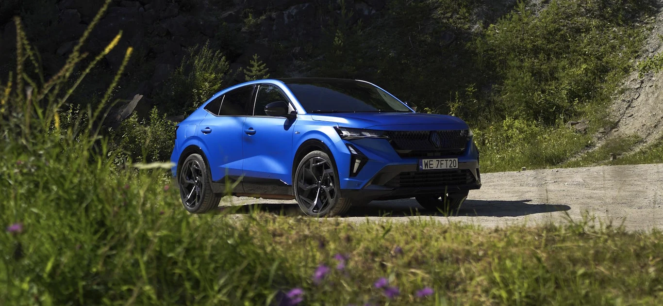 Renault Rafale to flagowy model w gamie francuskiego producenta Nowoczesny, niebieski samochód typu SUV zaparkowany na leśnej drodze, otoczony zielenią i dziką roślinnością, w tle widoczne drzewa oraz zbocze góry.