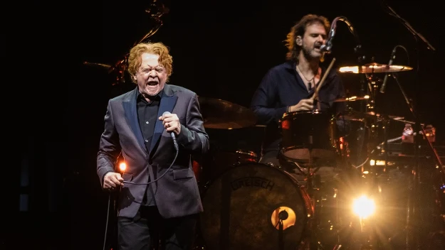 Wokalista w eleganckim garniturze śpiewa energicznie do mikrofonu na jasno oświetlonej scenie, za nim perkusista gra na perkusji na tle ciemnego tła z reflektorami skierowanymi na zespół.