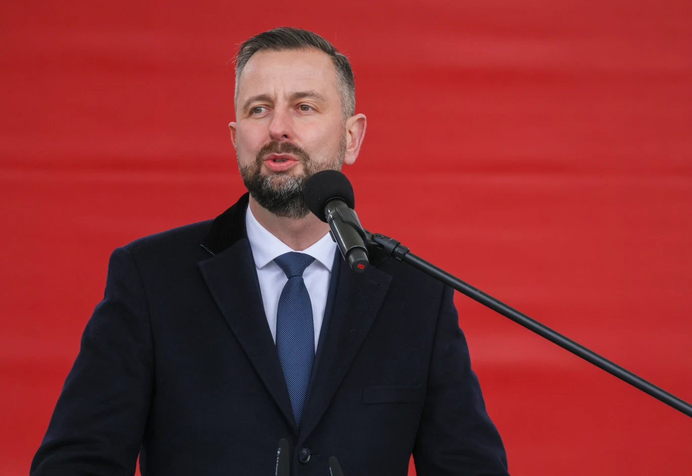 Szef MON o współpracy z Francją. "Polska dołącza do tego elitarnego grona" Mężczyzna w garniturze i krawacie przemawia do mikrofonu na tle jednolicie czerwonej ściany.