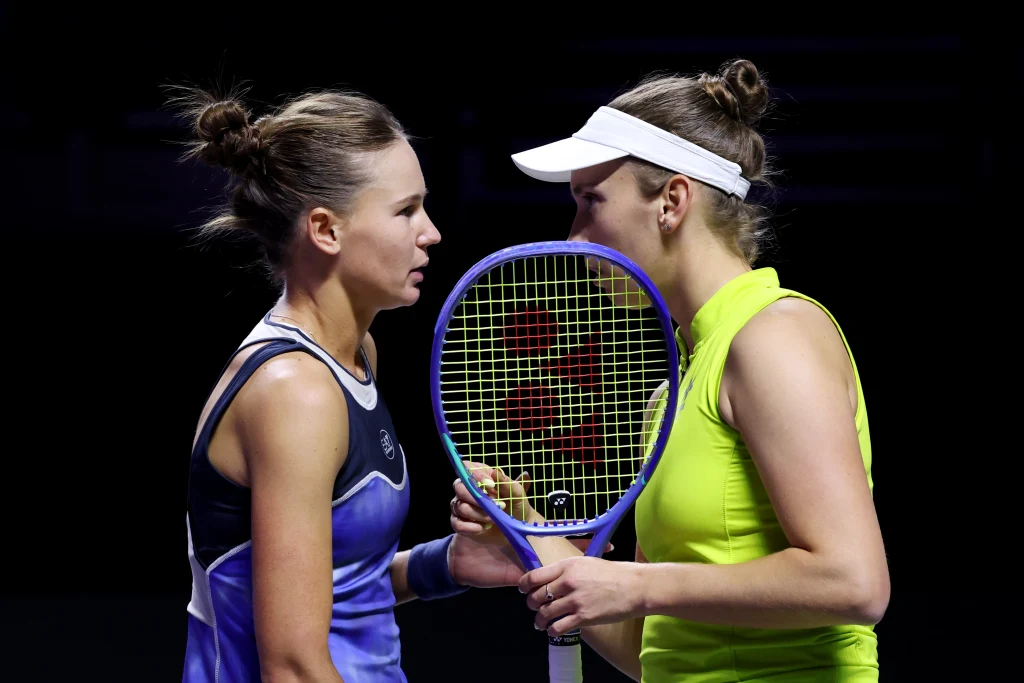Weronika Kudiermietowa (z lewej) i Elise Mertens Dwie tenisistki stoją twarzą w twarz przy siatce, jedna z nich trzyma rakietę tenisową i wydaje się rozmawiać z drugą. Obie zawodniczki są skoncentrowane, otoczenie jest ciemne, co podkreśla emocjonalny moment spotkania.