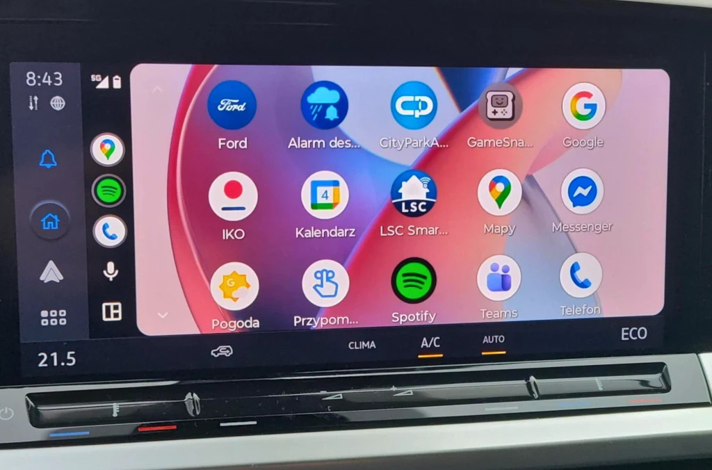Kolorowy ekran dotykowy samochodu z wyświetlonymi ikonami różnych aplikacji, takich jak mapy, muzyka, komunikatory, pogodynka i kalendarz. Poniżej ekranu widoczne są fizyczne przyciski panelu klimatyzacji.