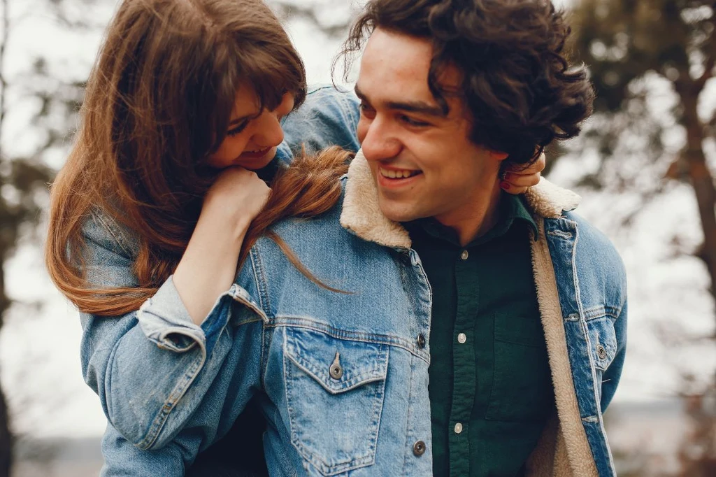Młoda para uśmiechnięta do siebie, kobieta obejmuje mężczyznę od tyłu, oboje ubrani w dżinsowe kurtki, w tle rozmyte drzewa.
