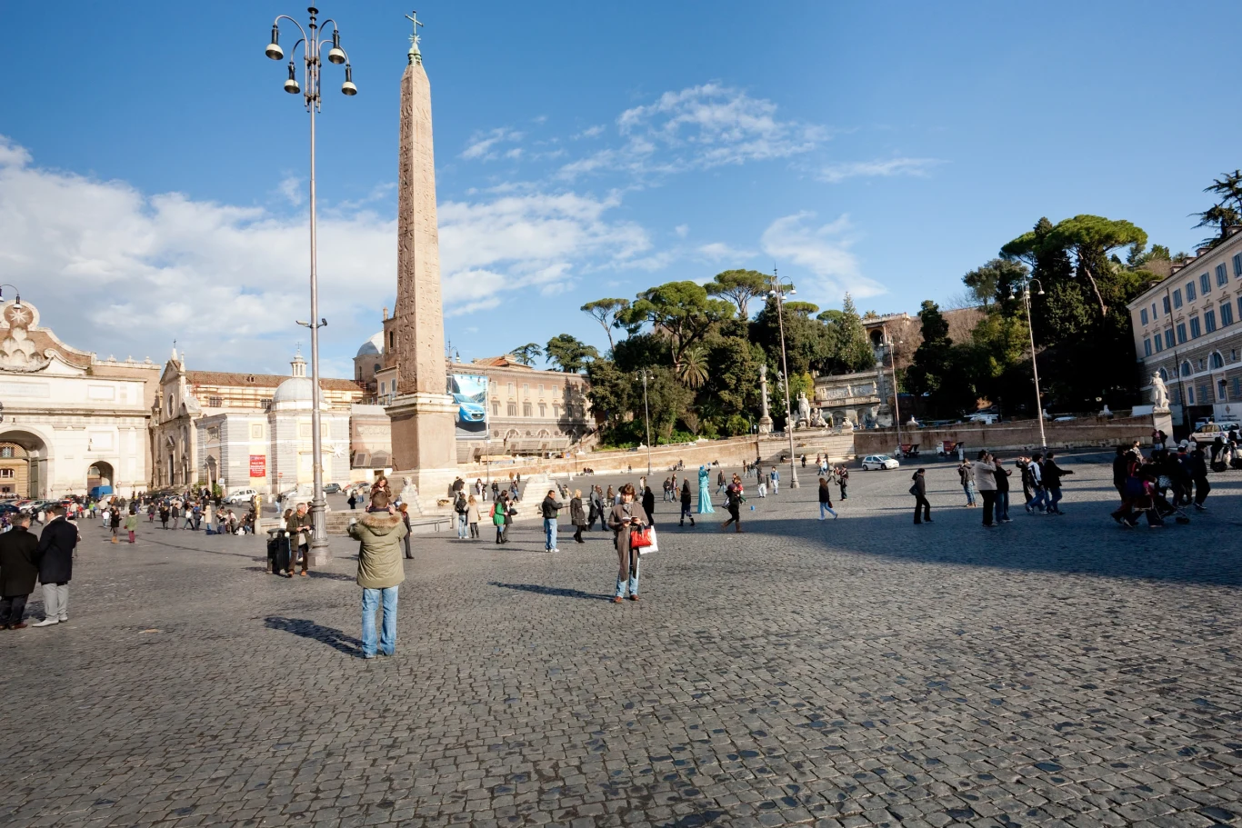 Rzym przyciąga turystów i kieszonkowców (zdj. ilustracyjne) Rozległy plac z brukowaną nawierzchnią, w centrum wysoki obelisk otoczony przez ludzi spacerujących pod otwartym niebem. Po bokach widoczne są zabytkowe budynki oraz drzewa i latarnie uliczne.