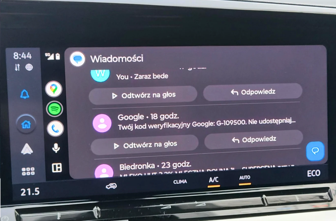 Ekran konsoli samochodowej z uruchomionym systemem Android Auto, na którym widoczne są powiadomienia z aplikacji wiadomości, w tym kod weryfikacyjny Google oraz komunikaty od użytkownika i sklepu Biedronka.