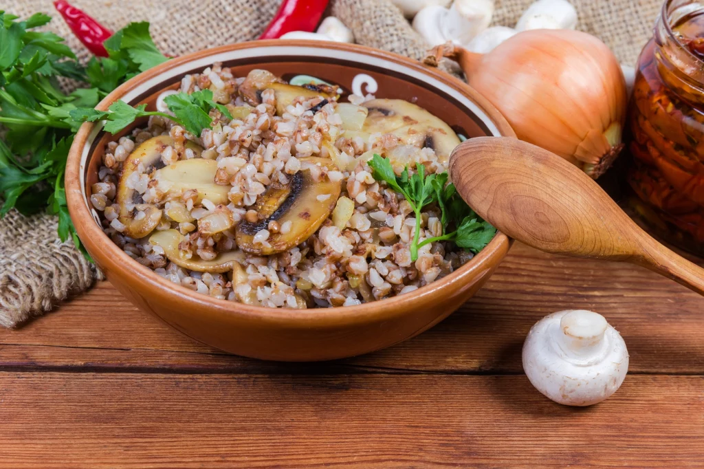 Kasza gryczana z duszonymi pieczarkami i cebulą w brązowej misce, ozdobiona świeżą natką pietruszki, w towarzystwie drewnianej łyżki oraz świeżych warzyw jak cebula, natka pietruszki i surowe pieczarki na rustykalnym drewnianym stole.