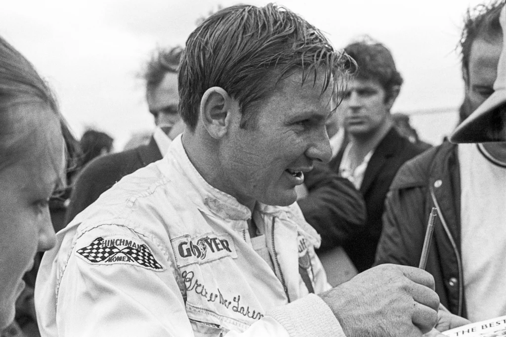 Bruce McLaren Kierowca wyścigowy podpisuje autograf, ubrany w biały kombinezon z naszywkami sponsorów, otoczony przez grupę ludzi skupionych wokół niego.