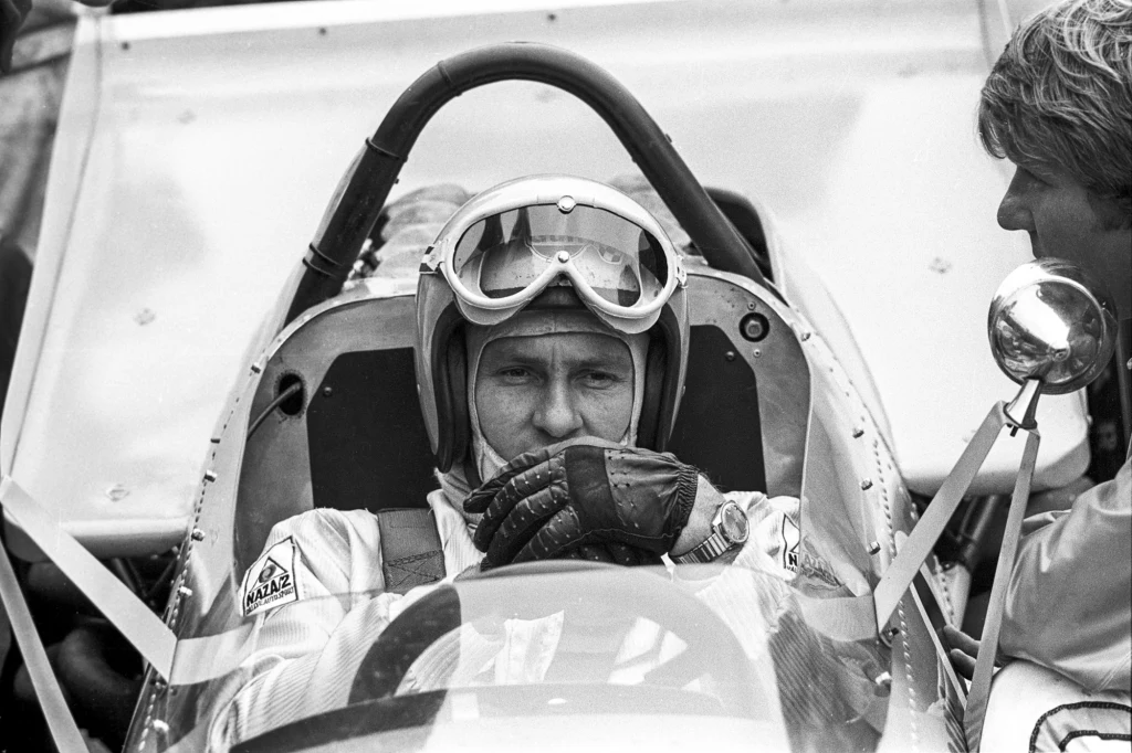 Bruce McLaren Kierowca wyścigowy w kombinezonie i kasku siedzi w kokpicie bolidu wyścigowego, zakładając rękawice, obok stoi inna osoba wpatrująca się w kierowcę.