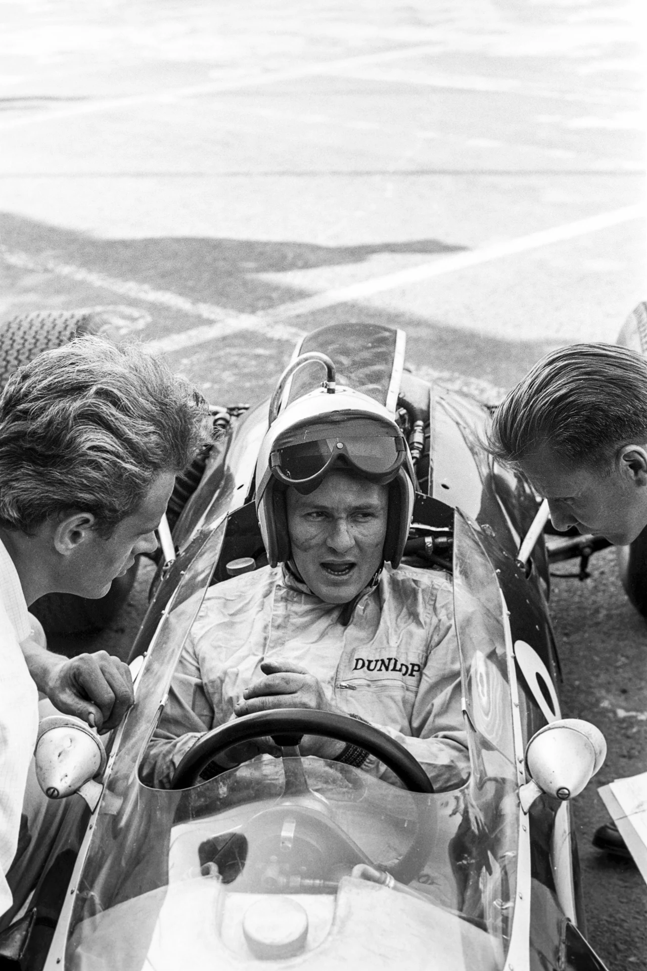 Bruce McLaren Kierowca wyścigowy siedzi w bolidzie, ubrany w kombinezon i kask ochronny z goglami, rozmawia z dwoma mężczyznami pochylonymi nad kokpitem. Widoczne szczegóły samochodu wyścigowego i elementy toru w tle.