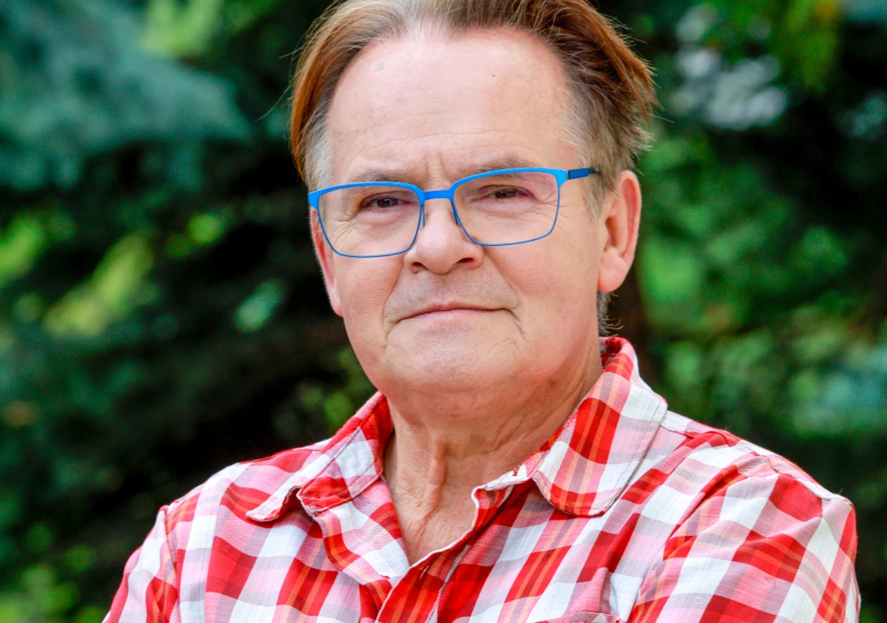 Piotr Pręgowski Starszy mężczyzna w okularach z niebieskimi oprawkami i koszuli w czerwoną kratę, stoi na tle zieleni, patrząc spokojnie w stronę aparatu