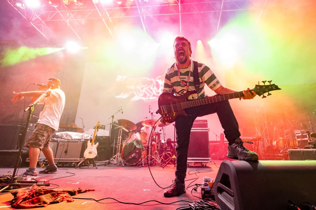 Zespół Maruja wydał album "Pain to Power" Energetyczny występ zespołu muzycznego na scenie z oświetleniem w odcieniach różu, zieleni i żółci, gitarzysta w centrum wyraża ekspresję twarzą i postawą, obok perkusja i drugi muzyk grający na trąbce.