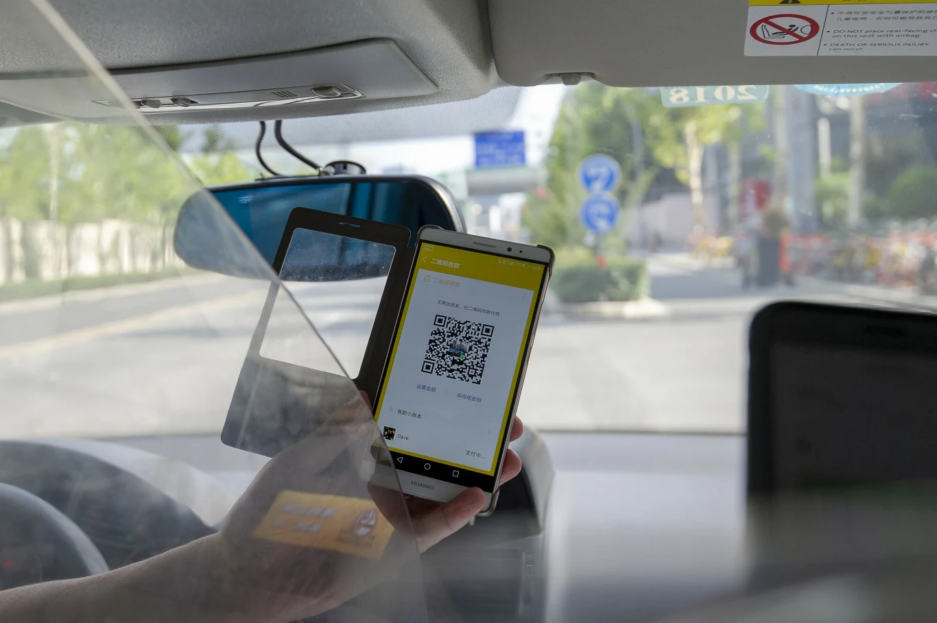 Ręka trzyma telefon wyświetlający kod QR w żółtej aplikacji, widoczne wnętrze samochodu oraz przezroczysta osłona oddzielająca kierowcę, za szybą widać ulicę z zielenią i znakami drogowymi.
