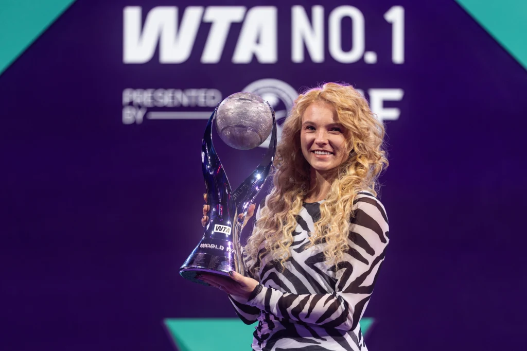 Katerina Siniakova z trofeum w Rijadzie Kobieta z długimi, kręconymi blond włosami trzyma w rękach duży puchar. W tle widoczny jest napis WTA No. 1 oraz logo WTA, a całość oświetla fioletowe światło podkreślające sportowy charakter sceny.