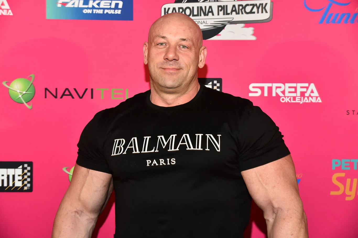 Mężczyzna o silnej posturze stoi na tle różowej ścianki z logotypami sponsorów, ubrany w czarną koszulkę Balmain Paris, uśmiecha się lekko do obiektywu.