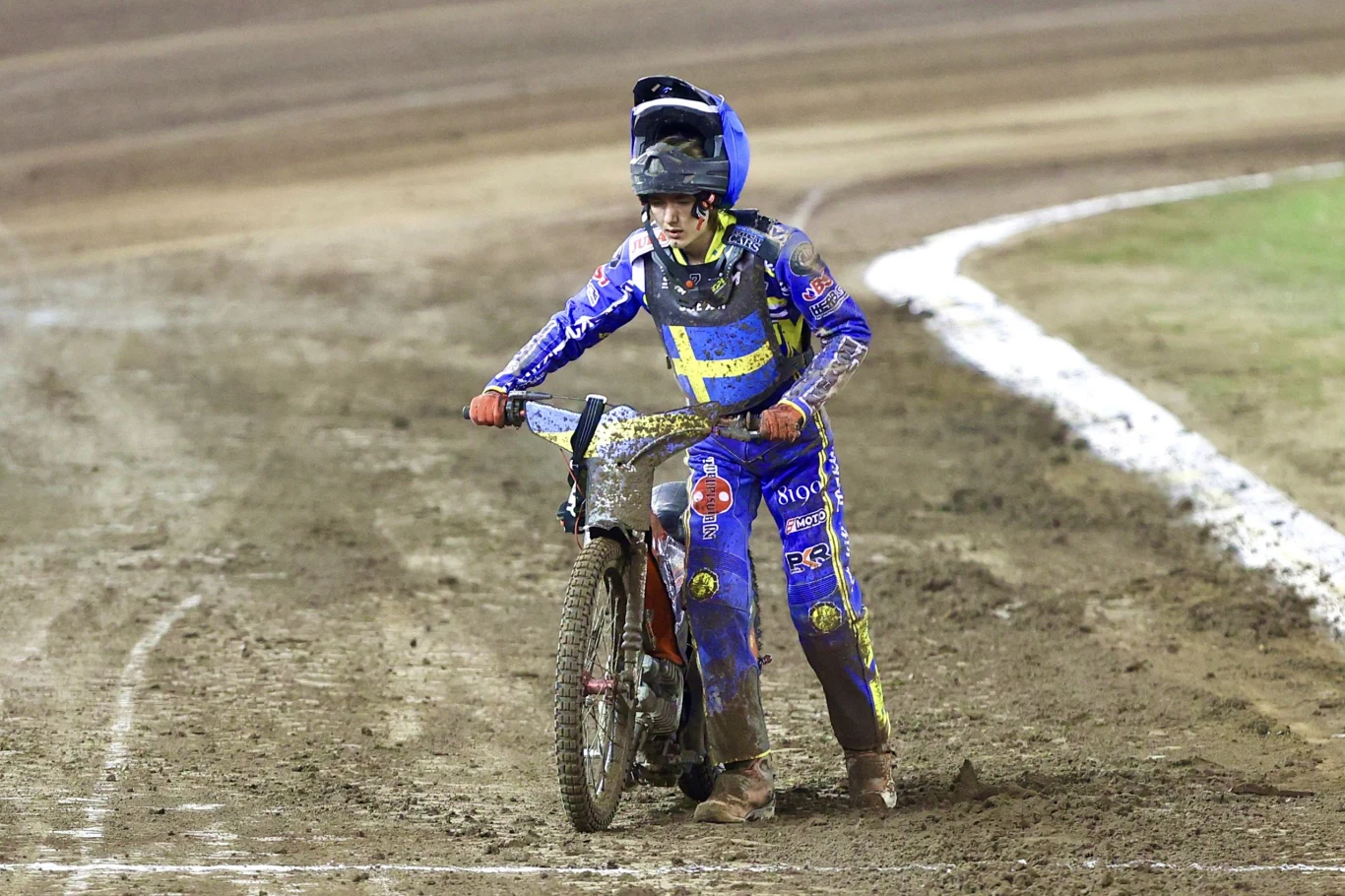 Zawodnik żużlowy w niebieskim kombinezonie i kasku prowadzi motocykl po torze pokrytym ziemią. Strój pokryty jest błotem, a tło to bieżnia i fragment trawnika.