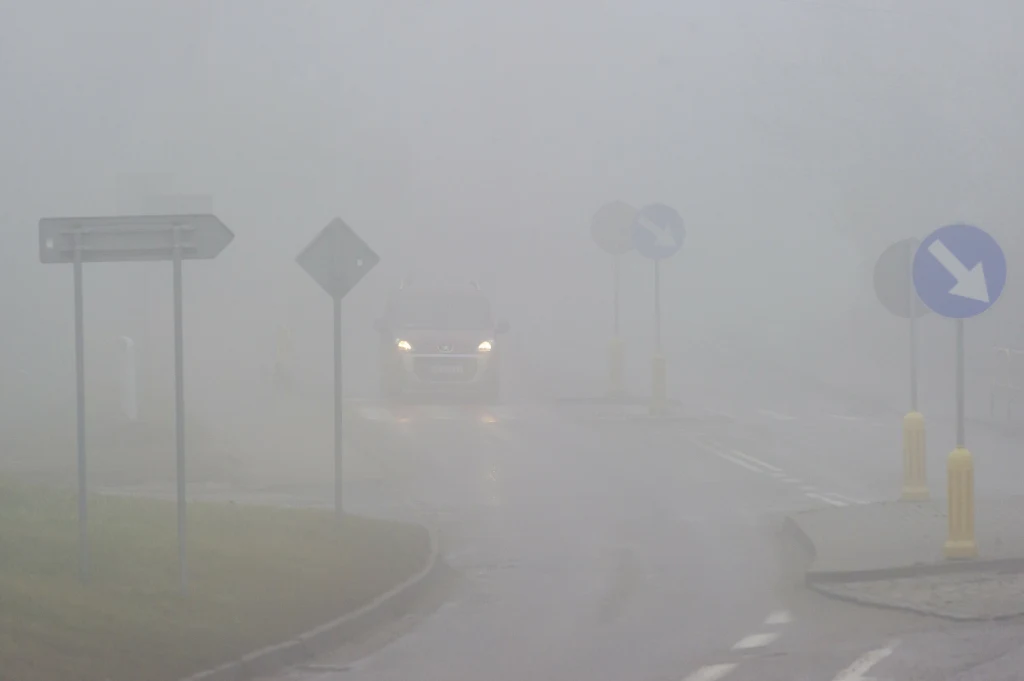IMGW wydał ostrzeżenia Gęsta mgła ograniczająca widoczność na drodze, ledwo widoczny samochód z włączonymi światłami mijania, znaki drogowe i wysepka rozdzielająca pasy ruchu.