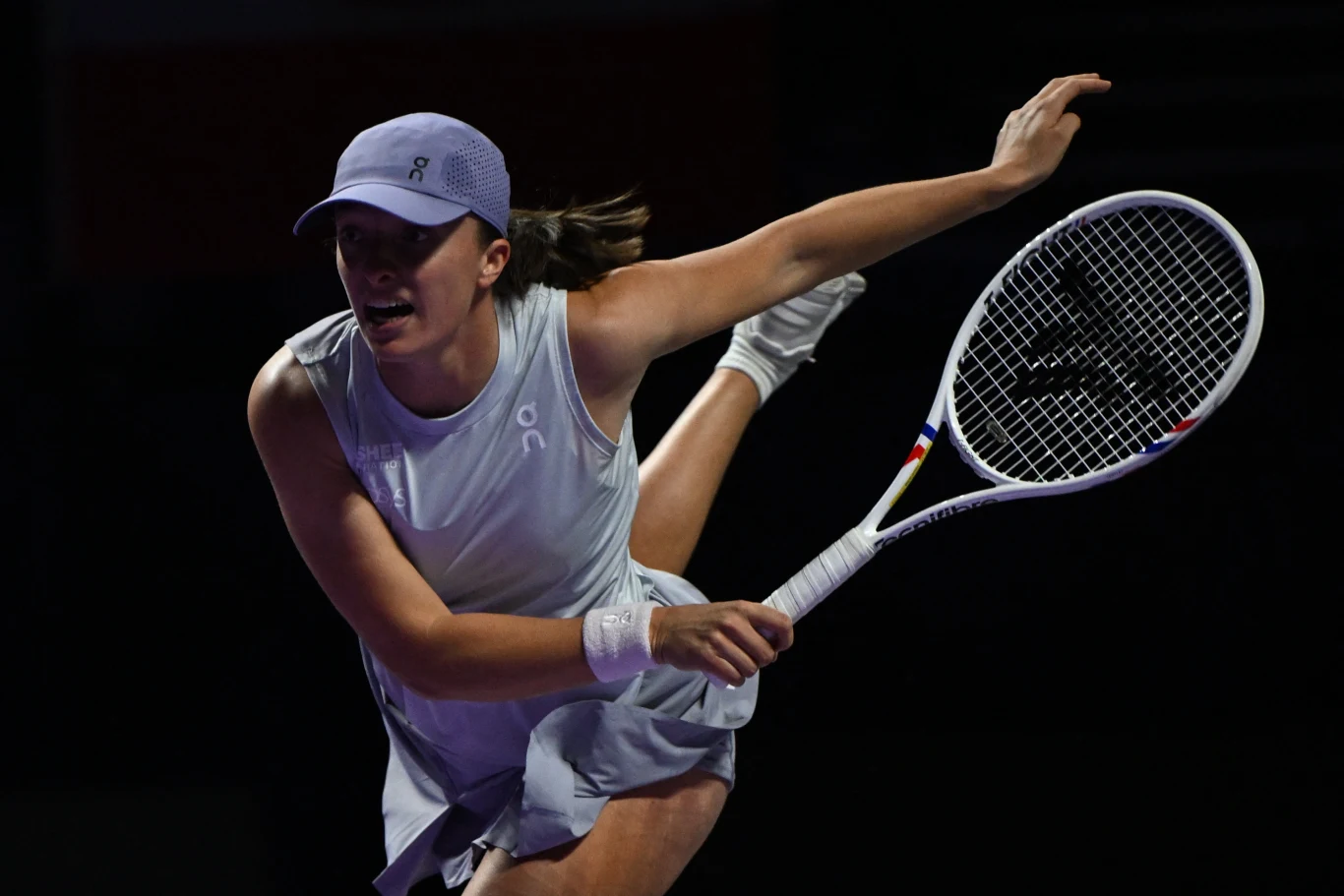 Iga Świątek rywalizowała z Amandą Anisimovą o awans do półfinału WTA Finals 2025 Tenisistka uderzająca rakietą w piłkę podczas intensywnej gry na korcie. W ciemnym tle wyraźnie widać ruch oraz wysiłek fizyczny zawodniczki ubraną w sportową sukienkę i czapkę.